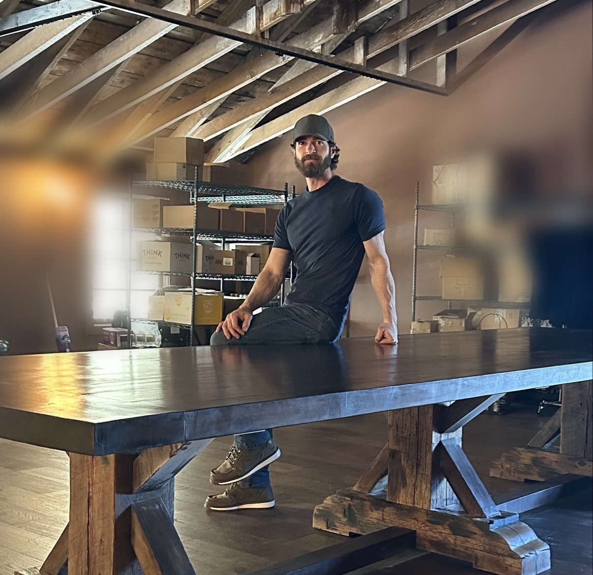 Adam Jessup seated on a large custom trestle table he built in his shop, beneath an exposed timber-frame ceiling
